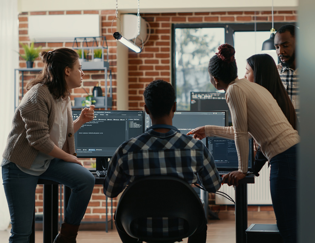 Four people in a modern office gather around a desk with computer monitors displaying code, collaborating on Azure Cloud Managed Services projects, with a brick wall and large windows in the background.