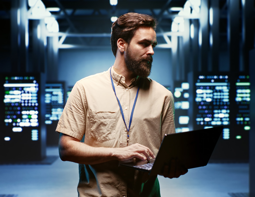 A man with a beard and short hair, wearing a beige shirt and lanyard, stands in a server room holding an open laptop, looking to the side—symbolizing expertise in Azure Managed Services with rows of servers visible in the background.