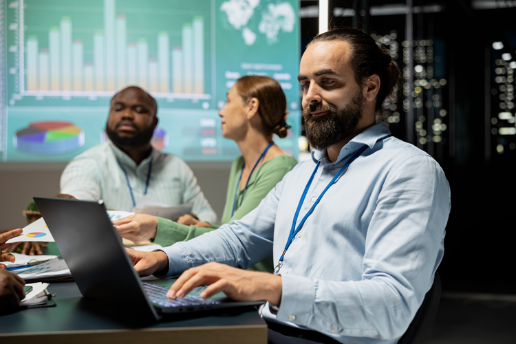 Three professionals sit at a table in a modern office, with one man typing on a laptop. Behind them, charts and graphs highlight Business Intelligence analytics as the group engages in a focused business meeting or discussion.