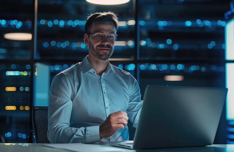 A man wearing glasses and a white shirt sits at a desk, focused on data security as he works on his laptop in a dimly lit office with large windows and city lights visible in the background.