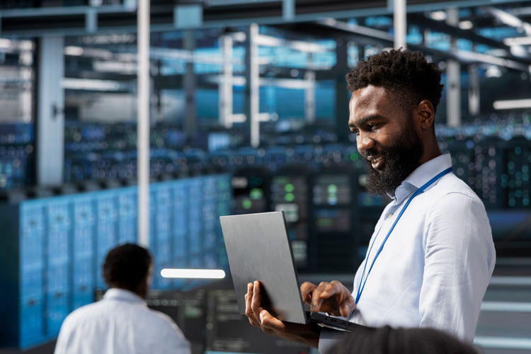 A man in a white shirt works on a laptop in a large server room filled with rows of blue computer servers, managing supported database platforms. Another person works in the background, highlighting the modern, high-tech environment.