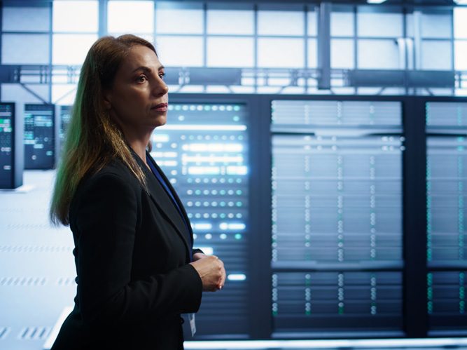 A woman in a business suit stands in a server room, surrounded by large racks of servers with glowing lights. She looks thoughtful, considering data analytics and visualization, with modern technology equipment in the background.