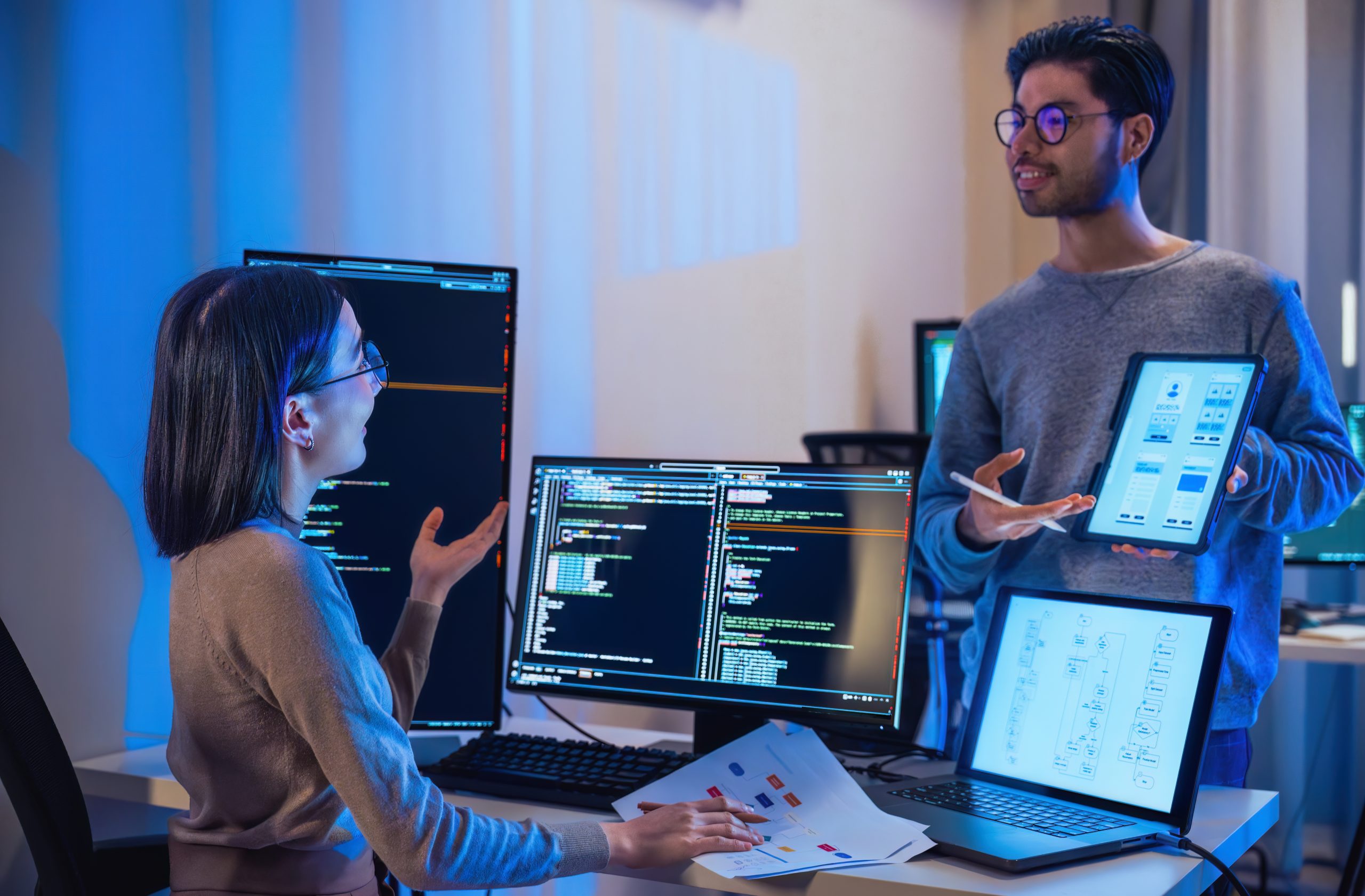 Two people in an office discuss software and platform support displayed on multiple monitors and an open laptop. One sits with printed documents while the other stands, holding a tablet showing a wireframe design. The room has a blue-lit ambiance.