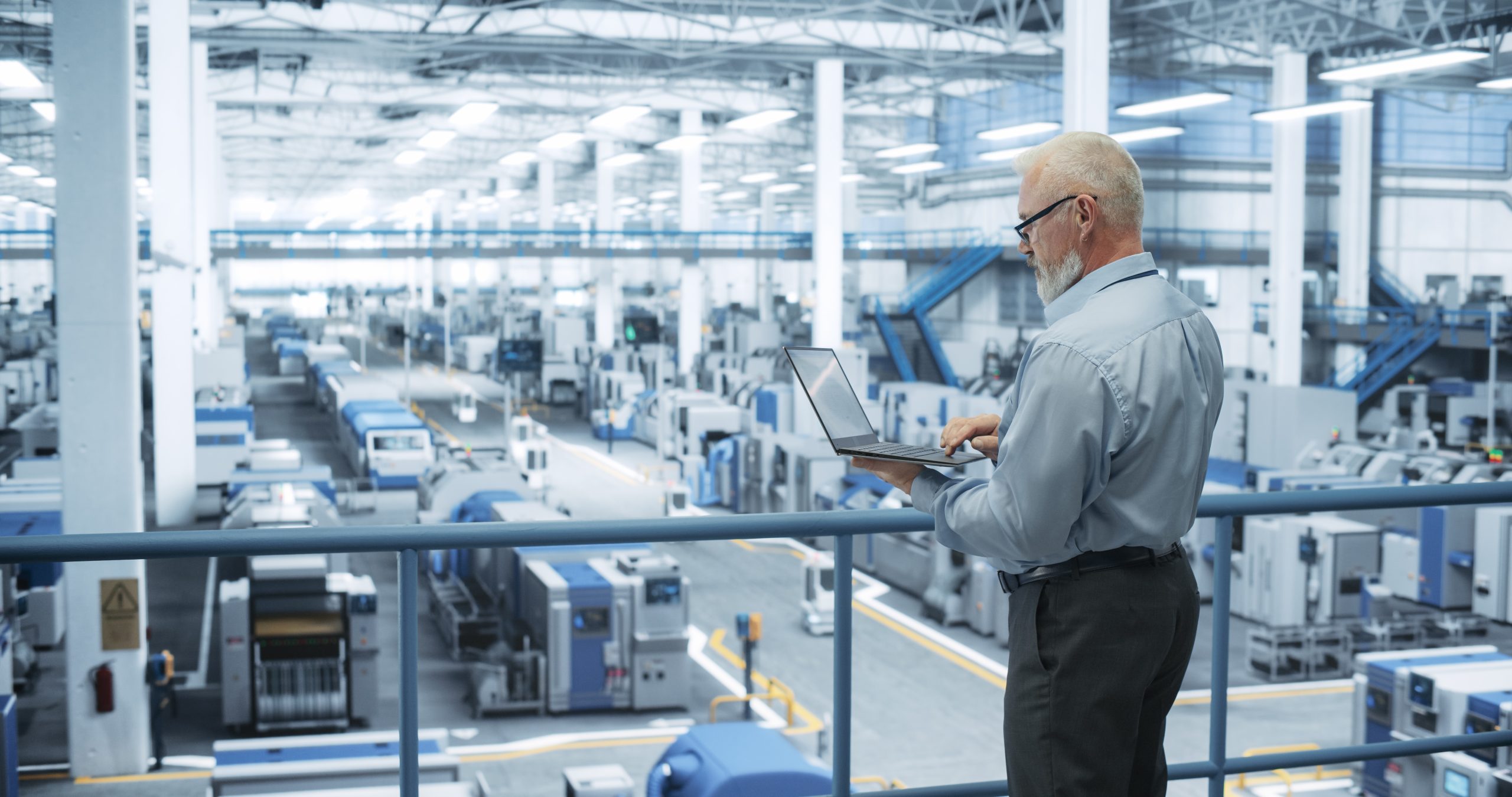 An older man in business attire stands on a balcony overlooking a large, modern factory floor filled with industrial machines, working on a laptop as he monitors the manufacturing process.