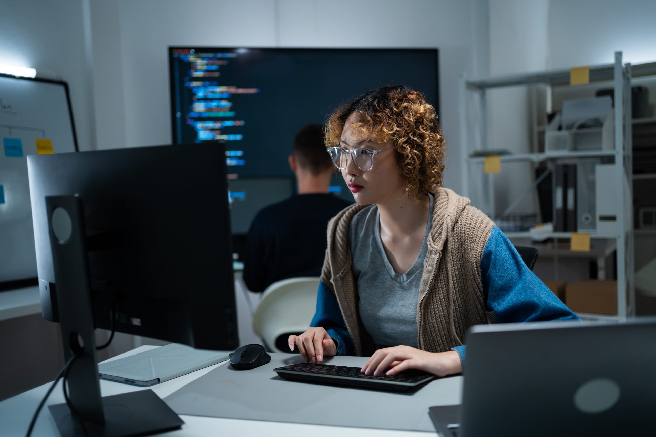 A person with curly hair and glasses works at a computer in an office, while another focuses on code displayed on a large monitor—both collaborating on hybrid cloud architecture solutions.