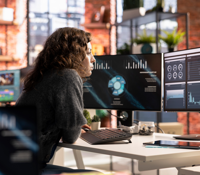 A woman sits at a desk in a modern office, exploring data visualizations and charts on multiple monitors powered by cloud services. The workspace is bright and filled with plants and natural light.
