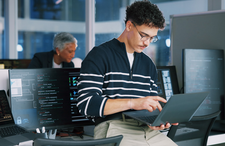 A young man in glasses uses a laptop while sitting at a desk in an office, with computer monitors displaying code and another person working in the background, conducting a Point in Time Assessment.