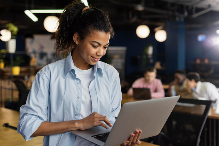 A woman stands in a modern office, smiling while using a laptop for a Point in Time Assessment. She wears a light blue shirt over a white top. In the background, several people are working at desks in the collaborative workspace.