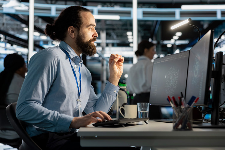 A man with a beard sits at a desk in an office, focused on a point in time assessment displayed across multiple computer monitors. Wearing a blue shirt and an ID badge, he works intently, with a mug and office supplies on the desk.