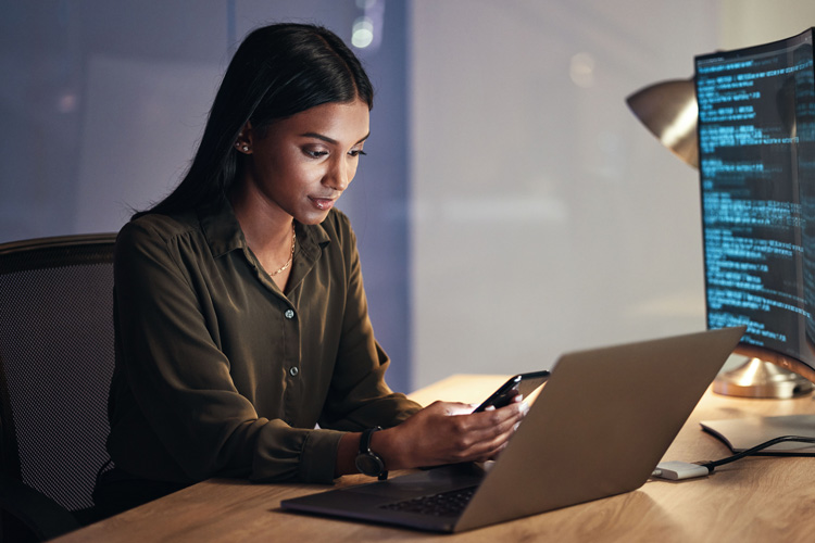 A woman sits at a desk with a laptop and a large monitor displaying backup code. She is looking at her phone and appears to be working in a modern office with soft lighting.