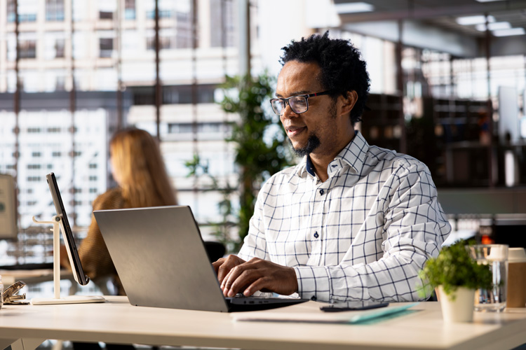 A man wearing glasses and a checkered shirt works on a laptop at a modern, bright office desk with plants and office supplies, focusing on BI Managed Services. A blurred colleague sits in the background.
