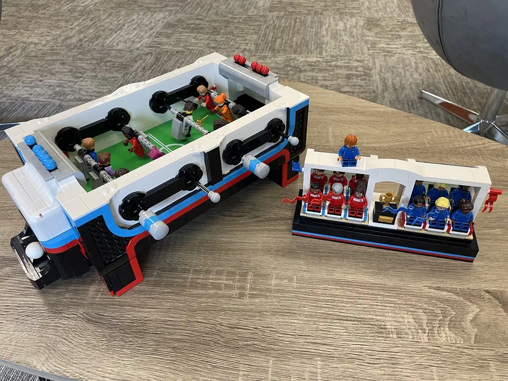 Lego foosball table with mini Lego players, next to a Lego model of stadium seating with Lego figures, both displayed on a wooden table with gray carpet in the background.