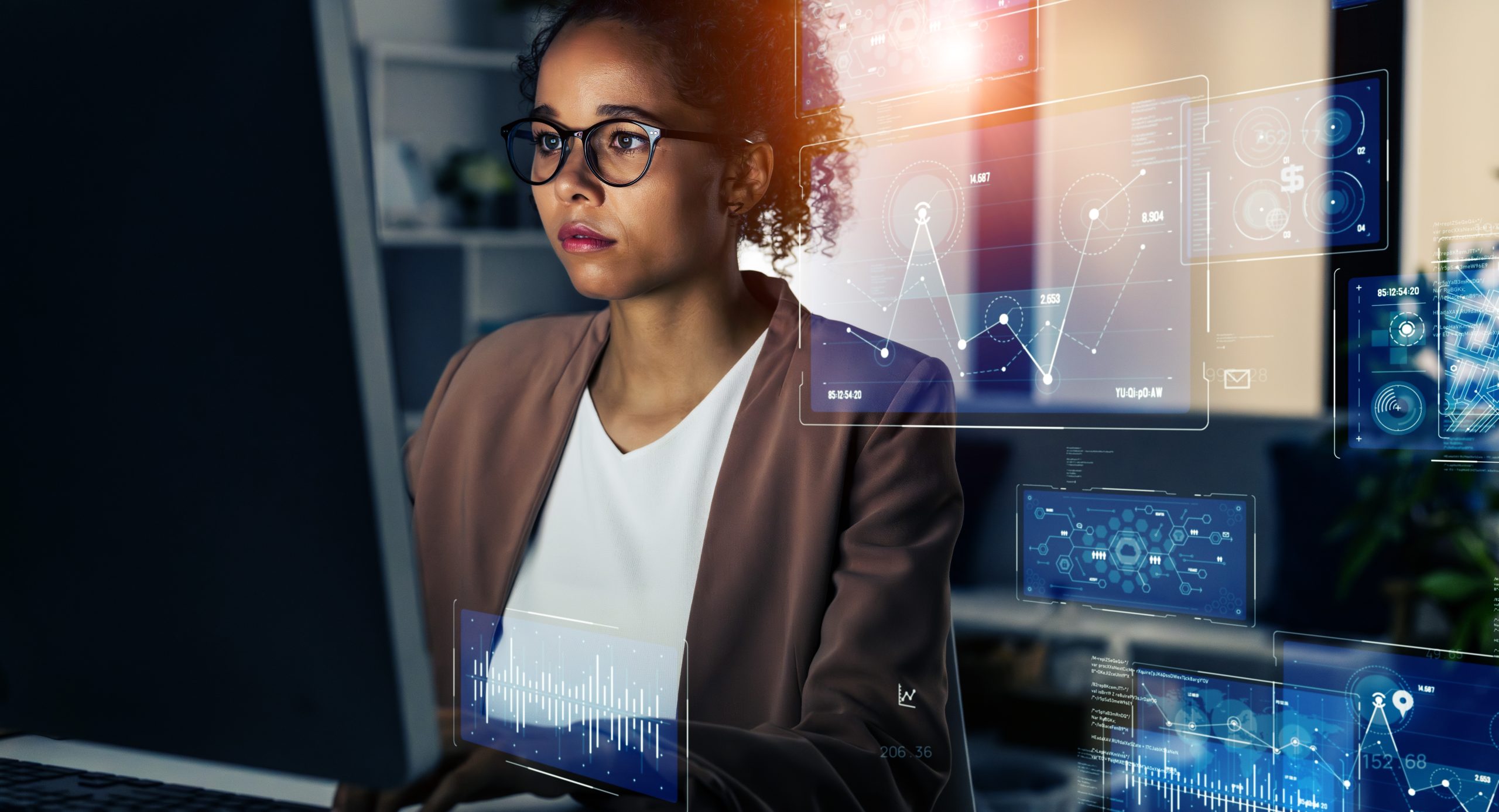 A woman wearing glasses and a blazer works at a computer, with transparent data charts projected in front of her, suggesting IT leadership or Fractional CIO services in a modern office setting.