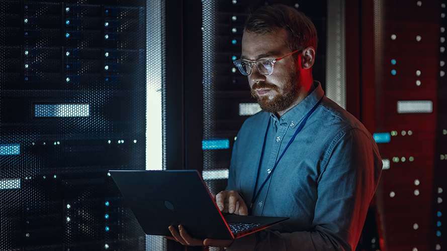 A man wearing glasses and a blue shirt works on a laptop in a dimly lit server room, surrounded by racks of servers with blue lights, focused on preventing database problems and maintaining smooth operations.